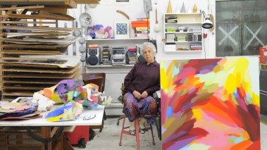 Artist Samia Halaby sitting on a chair in her studio next to a large abstract painting