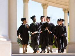 A group of joyous students in graduation gowns walk through a passage lined with pillars on a sunny day in university