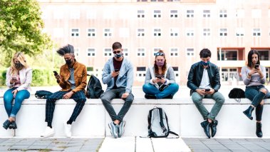 Friends using smart phone covered by mask on Covid third wave - Worried guys and girls watching video news on mobile smartphone - College students sitting at university campus