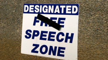 Sign reading "Designated Free Speech Zone" with "Free" crossed out.