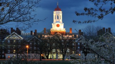 Harvard University at Night. Dunster House clock tower and red dome Illuminated at dusk, framed by cherry blossoms, blue sky and the banks of Charles River.